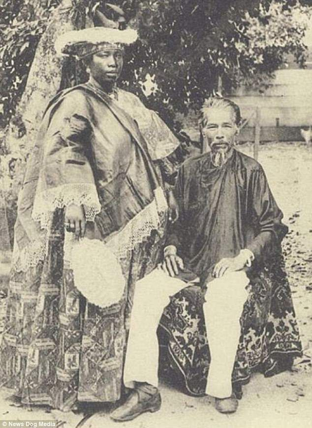 Um homem chinês senta-se ao lado de sua esposa em 1900. Ambos estão orgulhosos em roupas tradicionais
