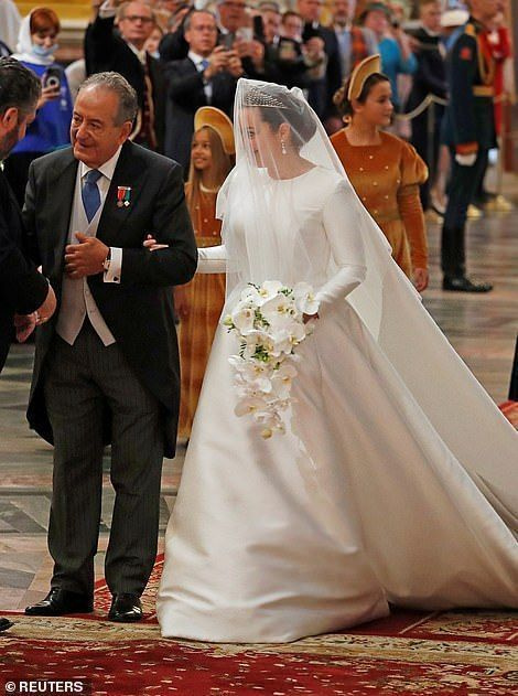 신부는 반짝이는 다이아몬드 티아라와 함께 하이넥, 긴소매, 부풀어오르는 스커트가 특징인 멋진 흰색 새틴 드레스를 입었습니다.